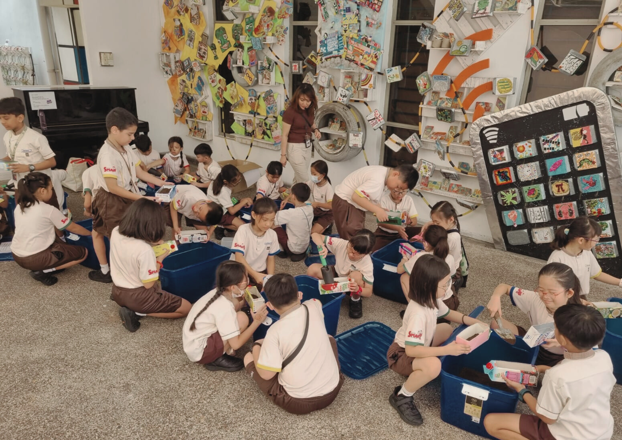 Schoolchildren in uniforms sort items into blue bins on the floor while a teacher watches by art-covered walls.